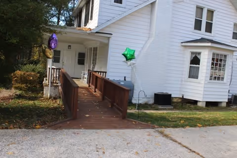 Exterior view of a white two-story house with a wooden ramp leading to the front porch. There are three balloons attached near the ramp, one purple, one gold, and one green. The house has multiple windows and is surrounded by grass and some bushes.