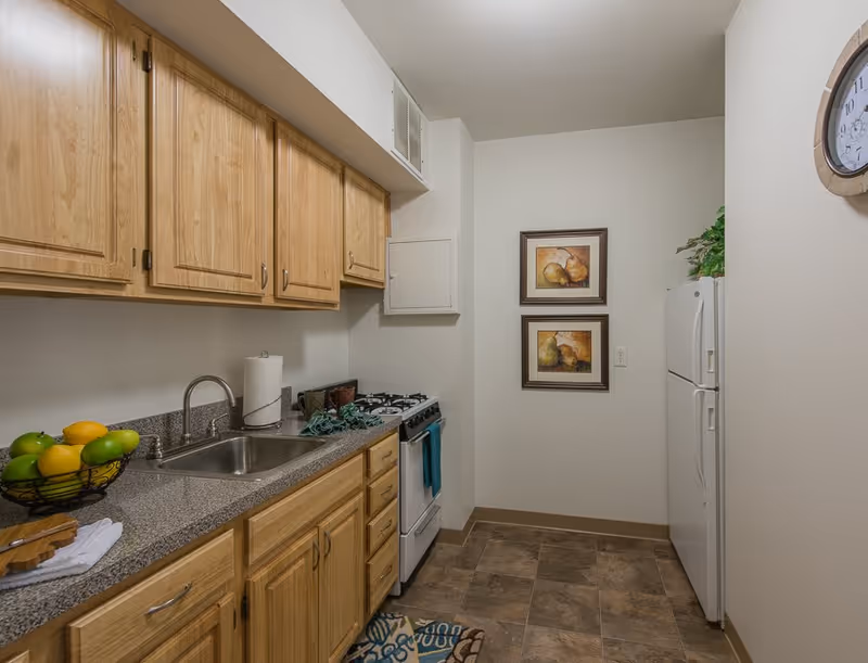 Narrow kitchen with wooden cabinets, a sink and countertop on the left, a stove and refrigerator on the right, and framed artwork on the back wall.