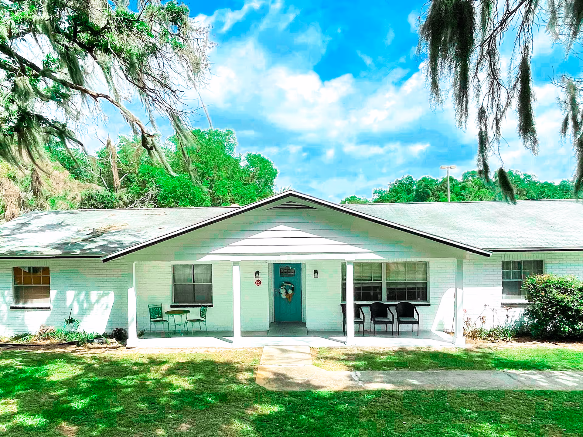 Front exterior view of a single-story white brick building with a teal front door decorated with a wreath. The building has a covered porch with two seating areas, one with a small table and two chairs, and the other with two black chairs. The lawn in front is green and shaded by large trees with hanging moss under a partly cloudy blue sky.