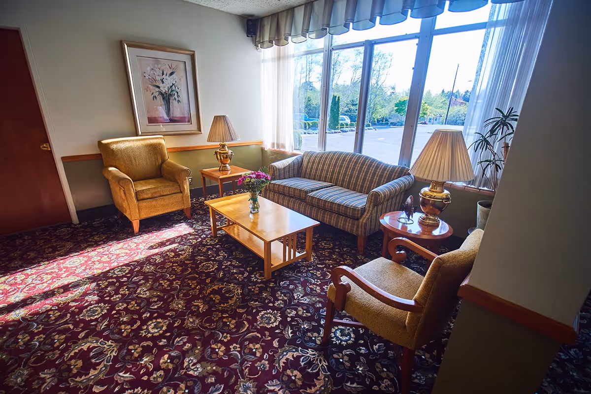 A cozy sitting area with a striped couch, two upholstered armchairs, a wooden coffee table with a vase of flowers, two side tables with lamps, a framed floral painting on the wall, and large windows letting in natural light overlooking a parking lot and trees outside.