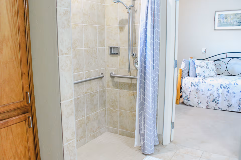 A tiled walk-in shower with grab bars and a handheld showerhead, partially covered by a gray and white patterned shower curtain. To the right, there is a view into a bedroom with a bed covered in floral bedding and a framed picture on the wall.