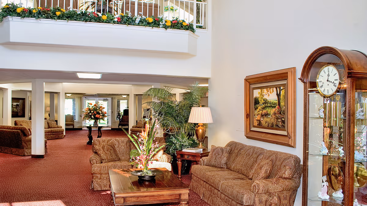 A spacious and well-lit living room area in a senior living facility with patterned brown sofas, a wooden coffee table with a floral arrangement, a side table with a lamp, a large grandfather clock, and a framed painting on the wall. There are plants and flowers decorating the space, with a balcony above adorned with a flower box.