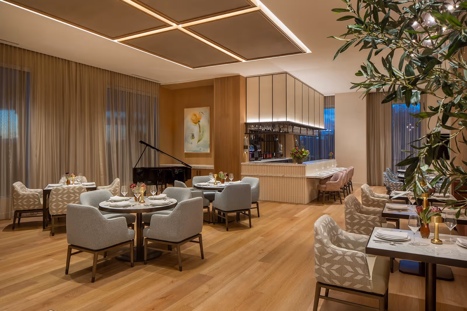 Elegant dining room with round and square tables, upholstered chairs, a bar area and a grand piano.