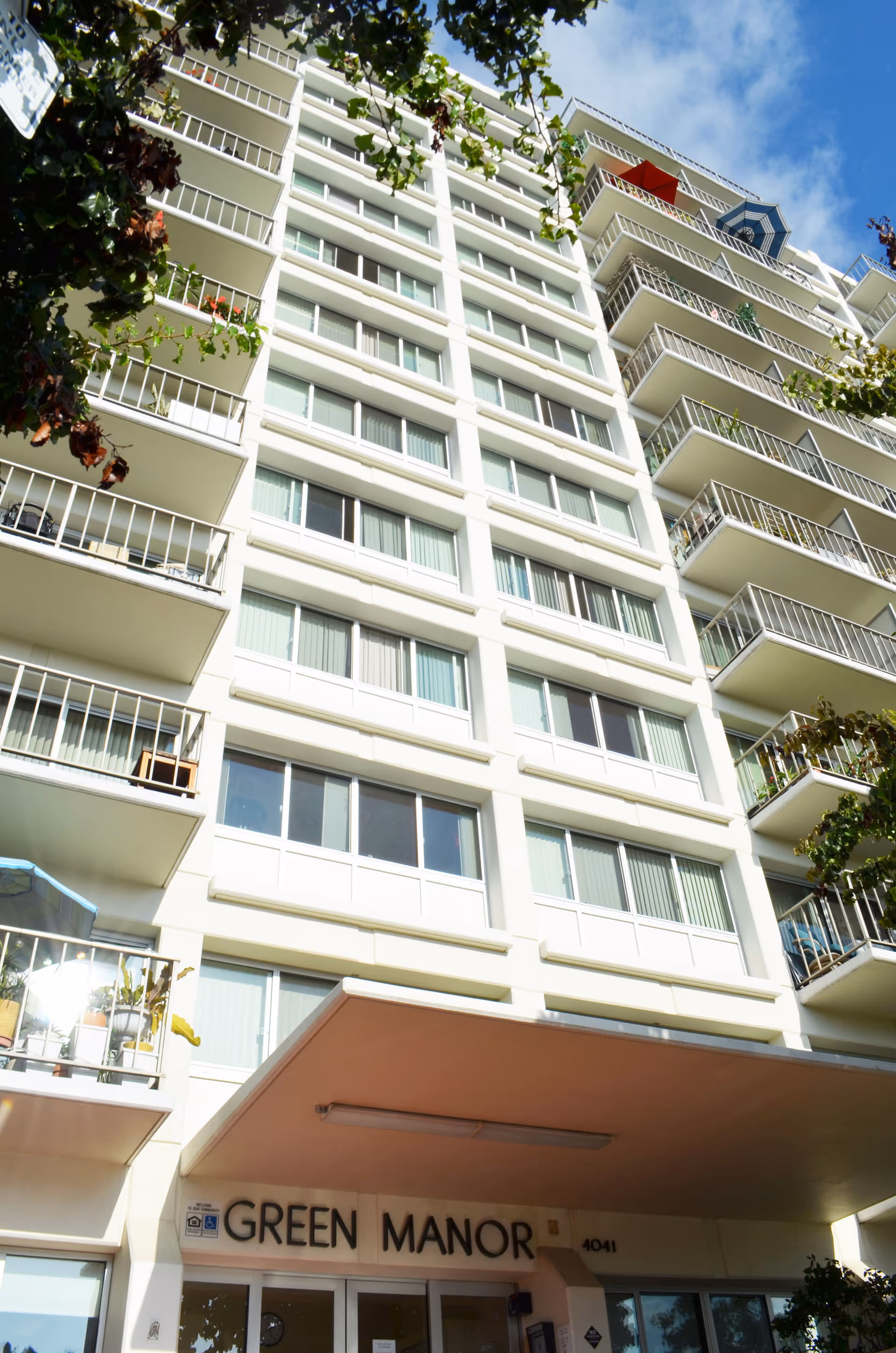 View looking up at the exterior of a multi-story residential building named Green Manor, showing balconies, windows, and some tree branches in the foreground under a partly cloudy sky.