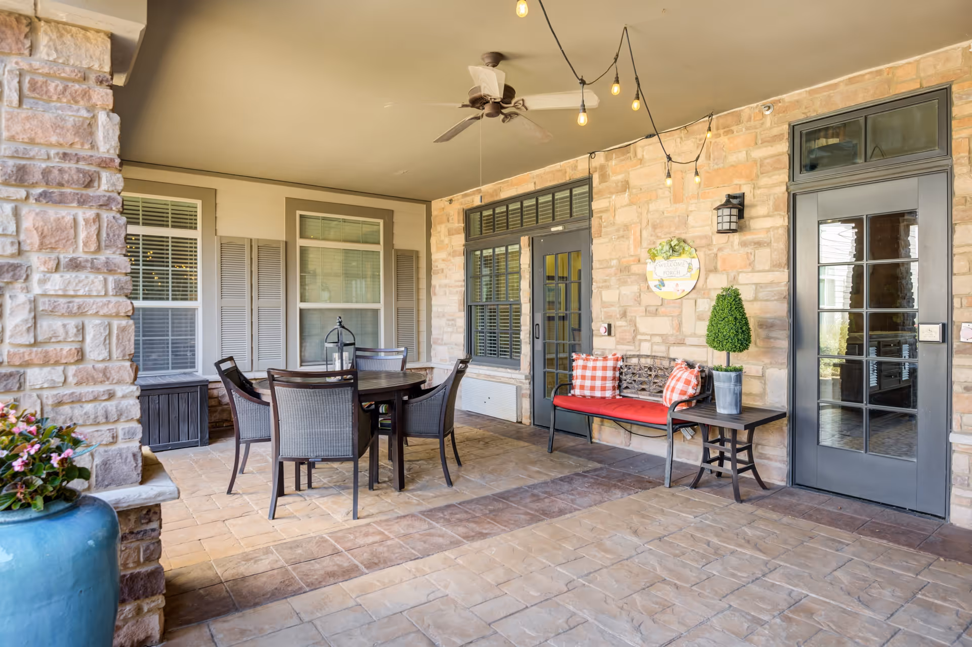 Covered outdoor patio area with stone walls and tiled floor, featuring a round table with four chairs, a bench with red cushions and checkered pillows, a small side table with a potted plant, string lights hanging from the ceiling, and two glass doors with window shutters.