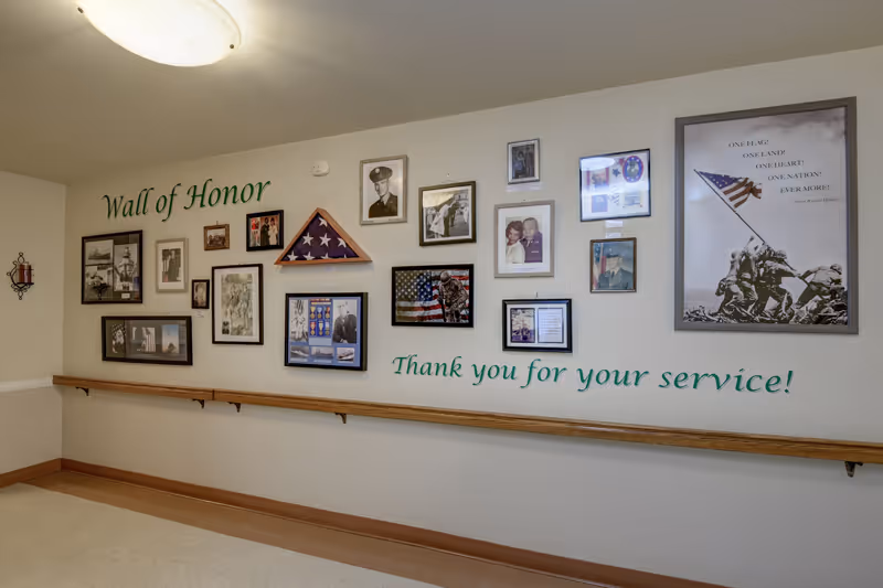 A hallway wall display titled 'Wall of Honor' featuring framed photographs, military memorabilia, and a folded American flag in a triangular case. Below the display, the text reads 'Thank you for your service!' A wooden handrail runs along the wall beneath the display.