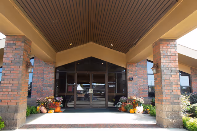 Entrance of a building with a covered porch supported by brick columns. The entrance has glass double doors with windows on either side. There are various potted plants and pumpkins arranged on both sides of the entrance, suggesting a seasonal decoration.