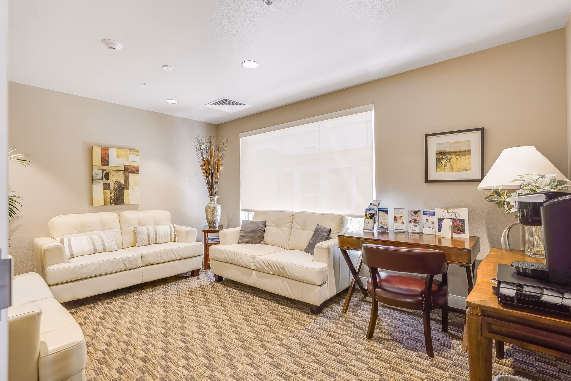 A cozy senior living common area with beige walls and carpeted floor. The room features two cream-colored leather sofas with decorative pillows, a wooden side table with a large silver vase holding dried branches, and a wooden desk with a chair. On the desk are several brochures and a table lamp with a white shade. A coffee maker and some binders are on a small wooden table next to the desk. A large window with a white roller shade allows natural light into the room. Two framed artworks hang on the walls.