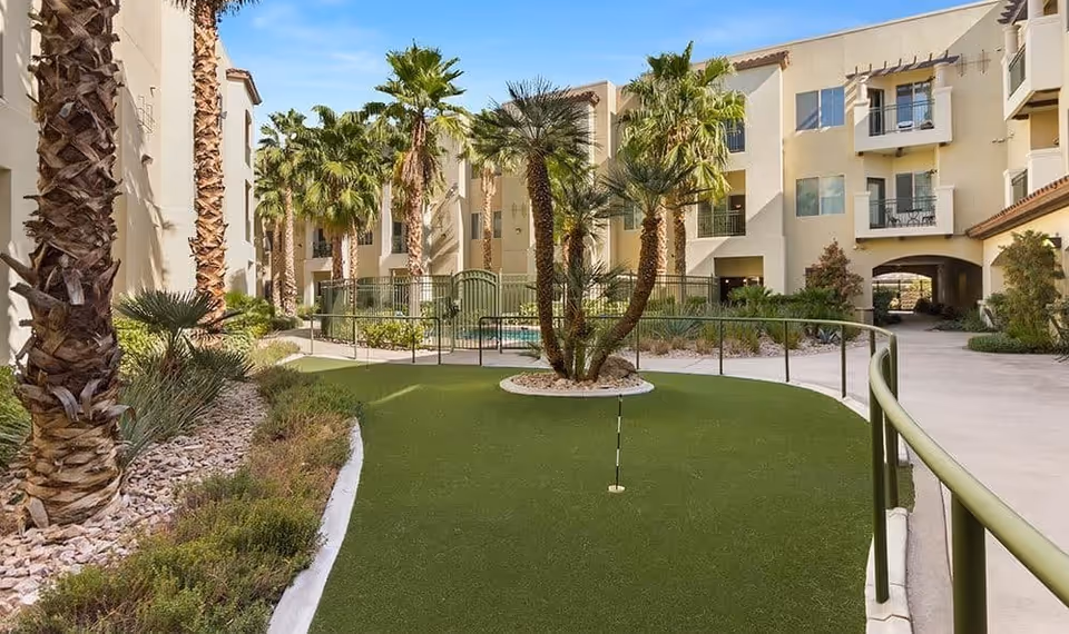 Outdoor courtyard area at Merrill Gardens at Green Valley Ranch featuring a putting green surrounded by palm trees and landscaped plants, with a multi-story residential building in the background under a clear blue sky.