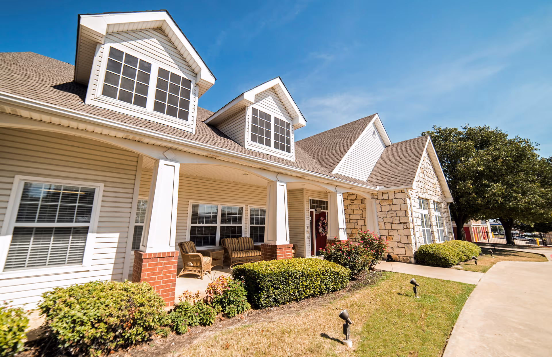 Front entrance of a single-story senior living building with a covered porch, outdoor seating, and landscaped bushes under a blue sky.
