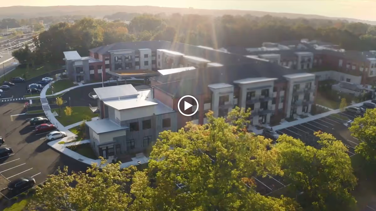 Aerial view of Brightview Wayne senior living facility showing a modern multi-story building surrounded by parking lots and green trees, with sunlight shining over the scene.
