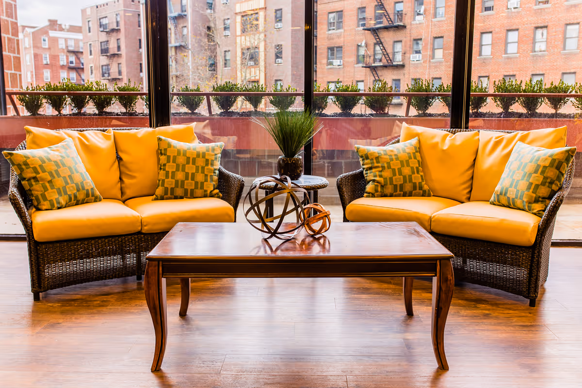 A cozy seating area with two wicker sofas featuring yellow cushions and patterned throw pillows, separated by a small round table with a potted plant. In front of the sofas is a wooden coffee table with decorative metal spheres. Large windows behind the seating area reveal an urban view with brick buildings and a row of small shrubs on the balcony.