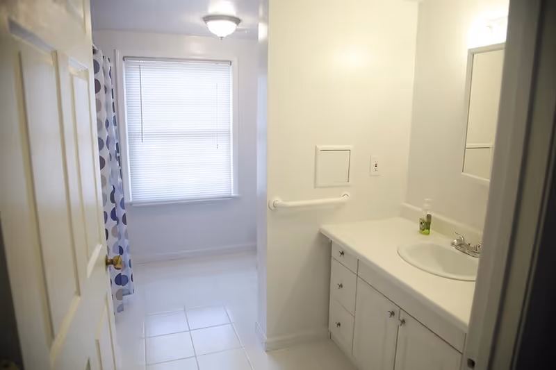 Bright bathroom with sink, mirror, shower curtain, and window in a senior living facility.
