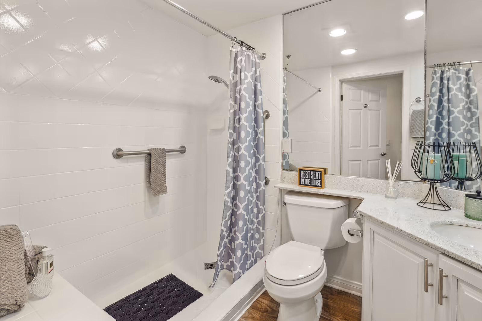 A clean bathroom with a white toilet, a countertop with a sink, and a large mirror above. There is a shower area with white tiled walls and a gray and white patterned shower curtain. A gray towel hangs on a metal bar inside the shower. On the countertop, there are decorative items including a small sign that reads 'BEST SEAT IN THE HOUSE', a reed diffuser, and candle holders. The floor is wood-style.