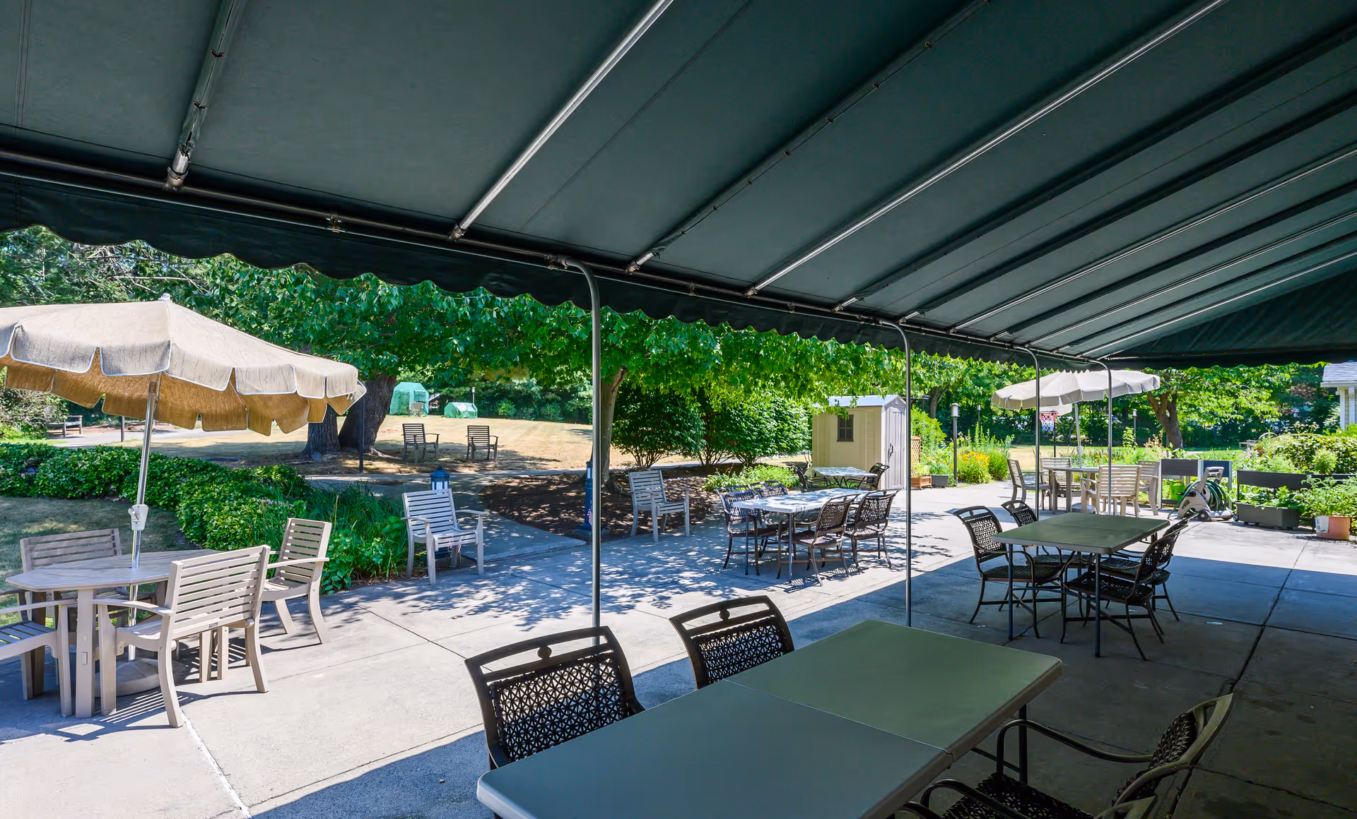 Outdoor patio area with multiple tables and chairs under a large green canopy. Additional tables with umbrellas are visible on the concrete patio. Surrounding the patio are green bushes, trees, and a grassy area with more seating and a small shed in the background.
