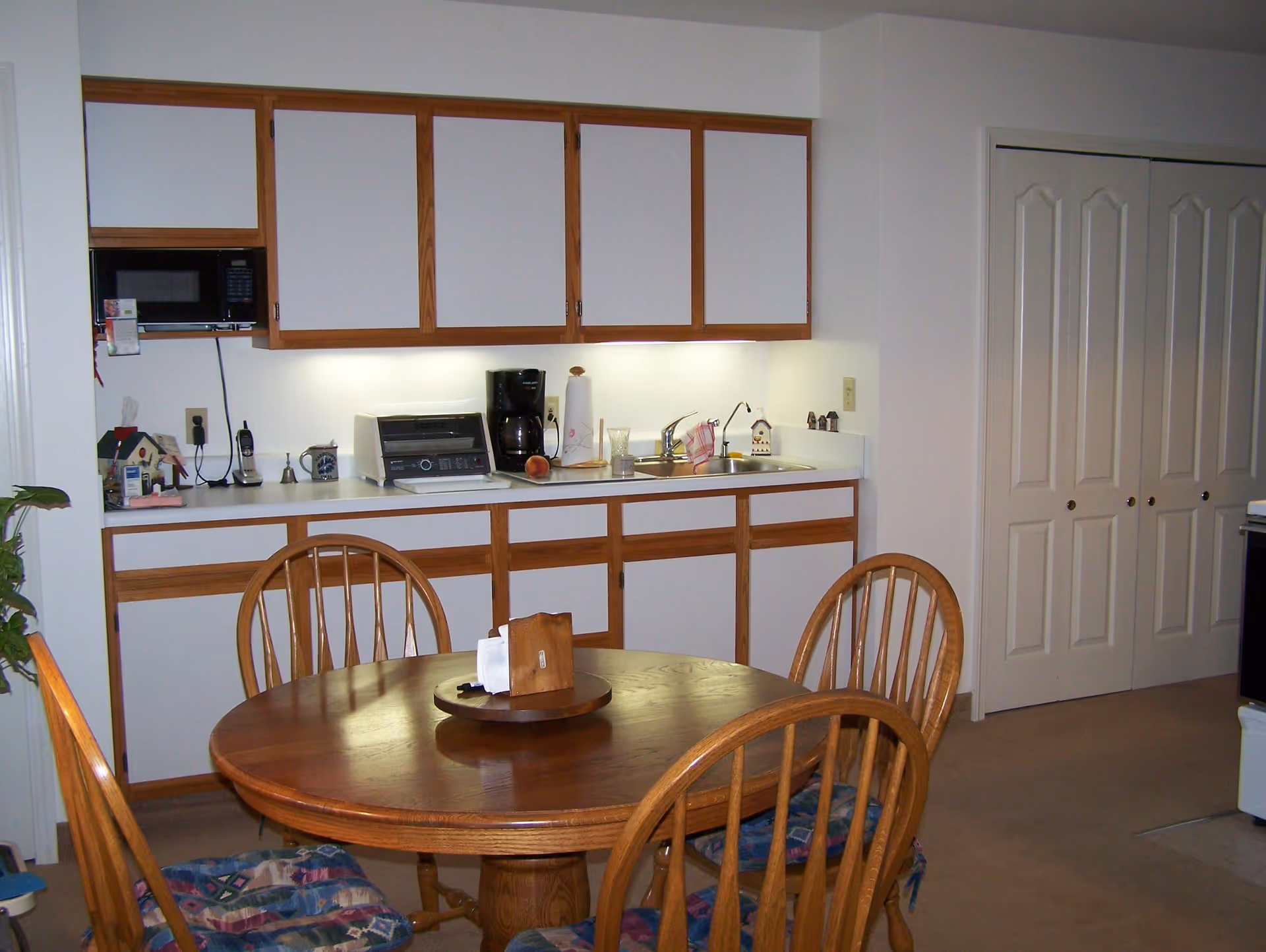 A small kitchen area with wooden cabinets featuring white doors, a countertop with a coffee maker, toaster oven, paper towel holder, and a sink. In front of the kitchen is a round wooden dining table with four wooden chairs, each with patterned seat cushions. There is a double-door closet to the right and a plant partially visible on the left.