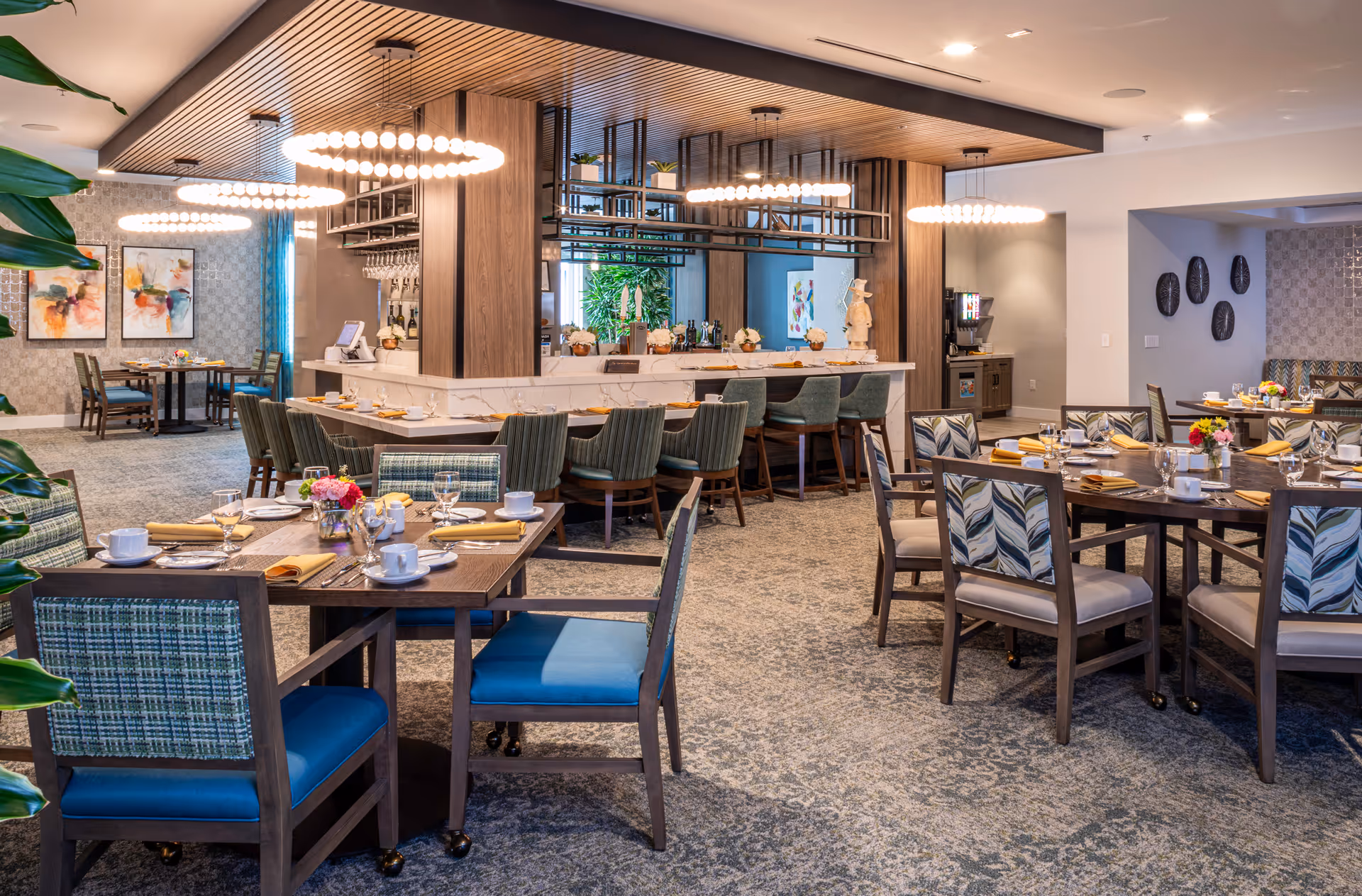 A spacious and well-lit dining room with multiple tables set with plates, cups, glasses, and yellow napkins. The room features modern circular pendant lights hanging from a wooden slatted ceiling above a bar area with high chairs. The walls are decorated with framed artwork and decorative plates, and there are plants adding greenery to the space.