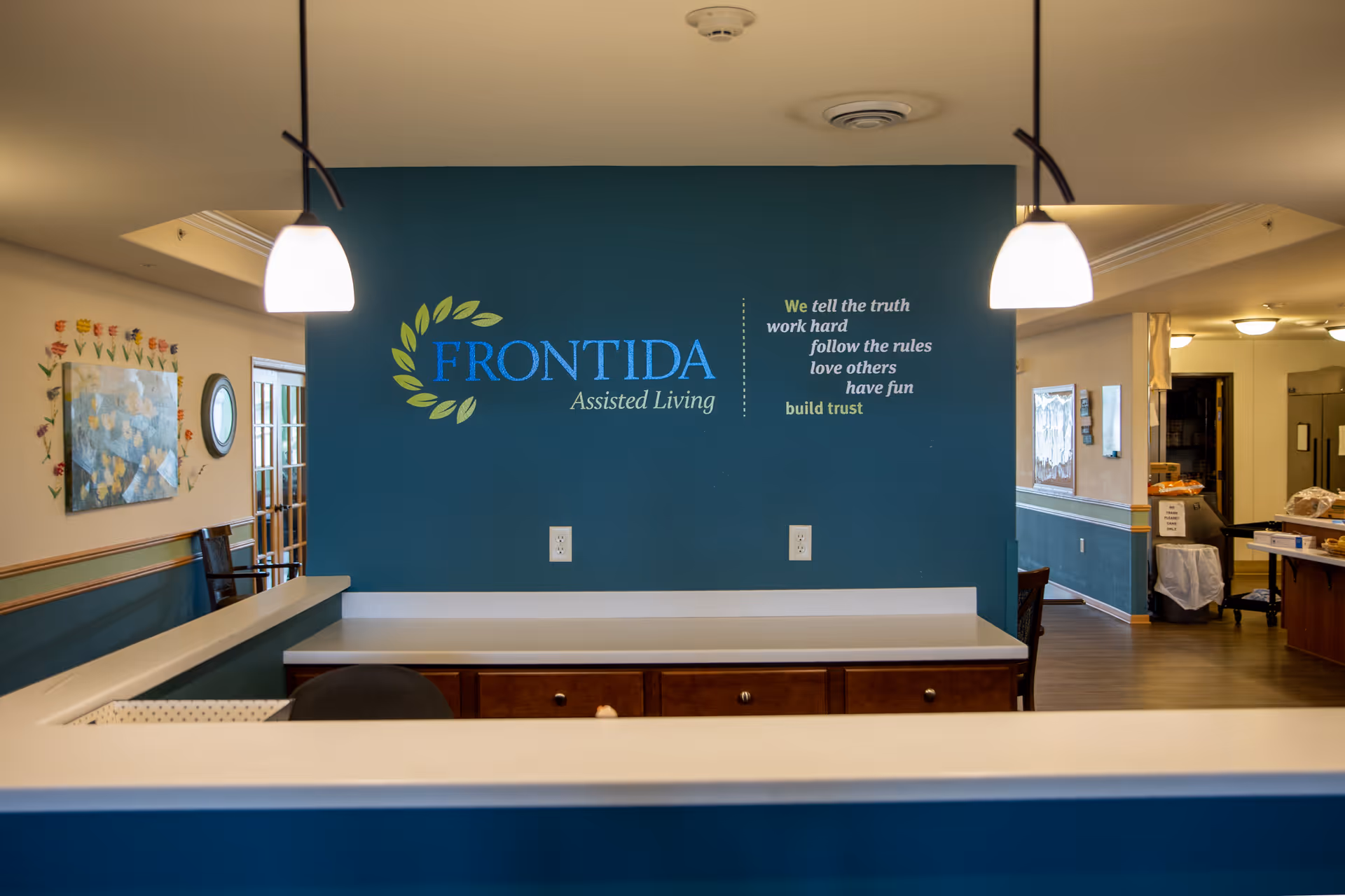 Reception area of Frontida Assisted Living with a blue accent wall displaying the facility's name and values. The counter is white with wooden drawers underneath, and two pendant lights hang from the ceiling. The surrounding area includes a hallway and some wall decorations.