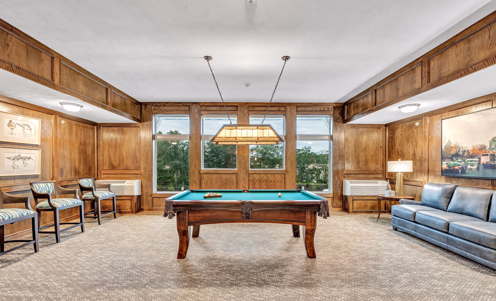A well-lit recreational room with a pool table in the center, surrounded by wooden panel walls. There are three striped cushioned chairs on the left side, a blue leather sofa on the right, and large windows at the back showing greenery outside. A rectangular hanging light fixture is above the pool table.