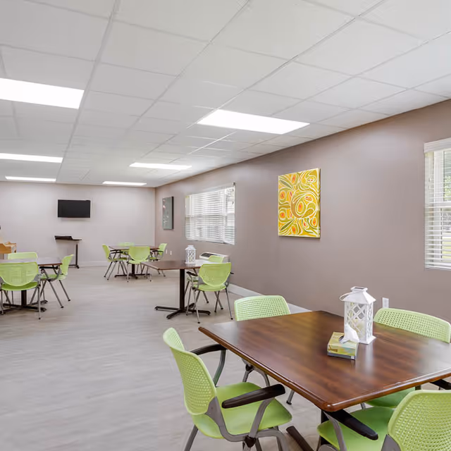 Bright communal dining/activity room with wooden tables, lime-green chairs, wall art, windows, and a wall-mounted TV.