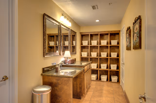 Interior view of a well-lit bathroom with a double sink vanity featuring granite countertops and wooden cabinets. Above the sinks are two large framed mirrors and a light fixture with three bulbs. To the right, there is a wooden shelving unit with multiple cubbies, each holding a white storage basket. The walls are painted beige, and there are two decorative floral paintings on the right wall. The floor is tiled, and a silver trash can is placed near the vanity.