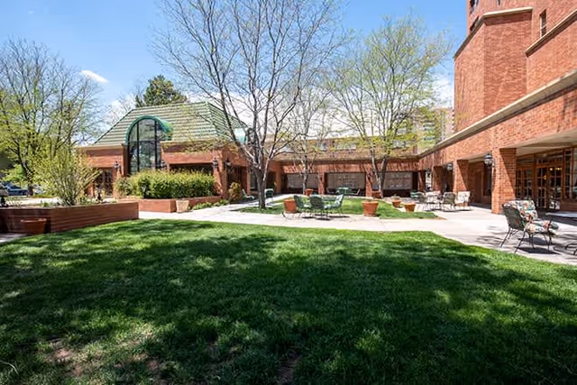 Outdoor courtyard area at Brookdale Parkplace featuring green grass, leafless and leafy trees, several patio chairs and tables, and red brick buildings surrounding the space under a clear blue sky.