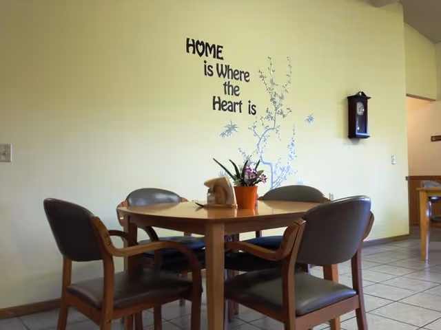Small dining area with a round wooden table and four chairs, a potted plant centerpiece, a wall clock, and a wall decal reading "Home is Where the Heart is."
