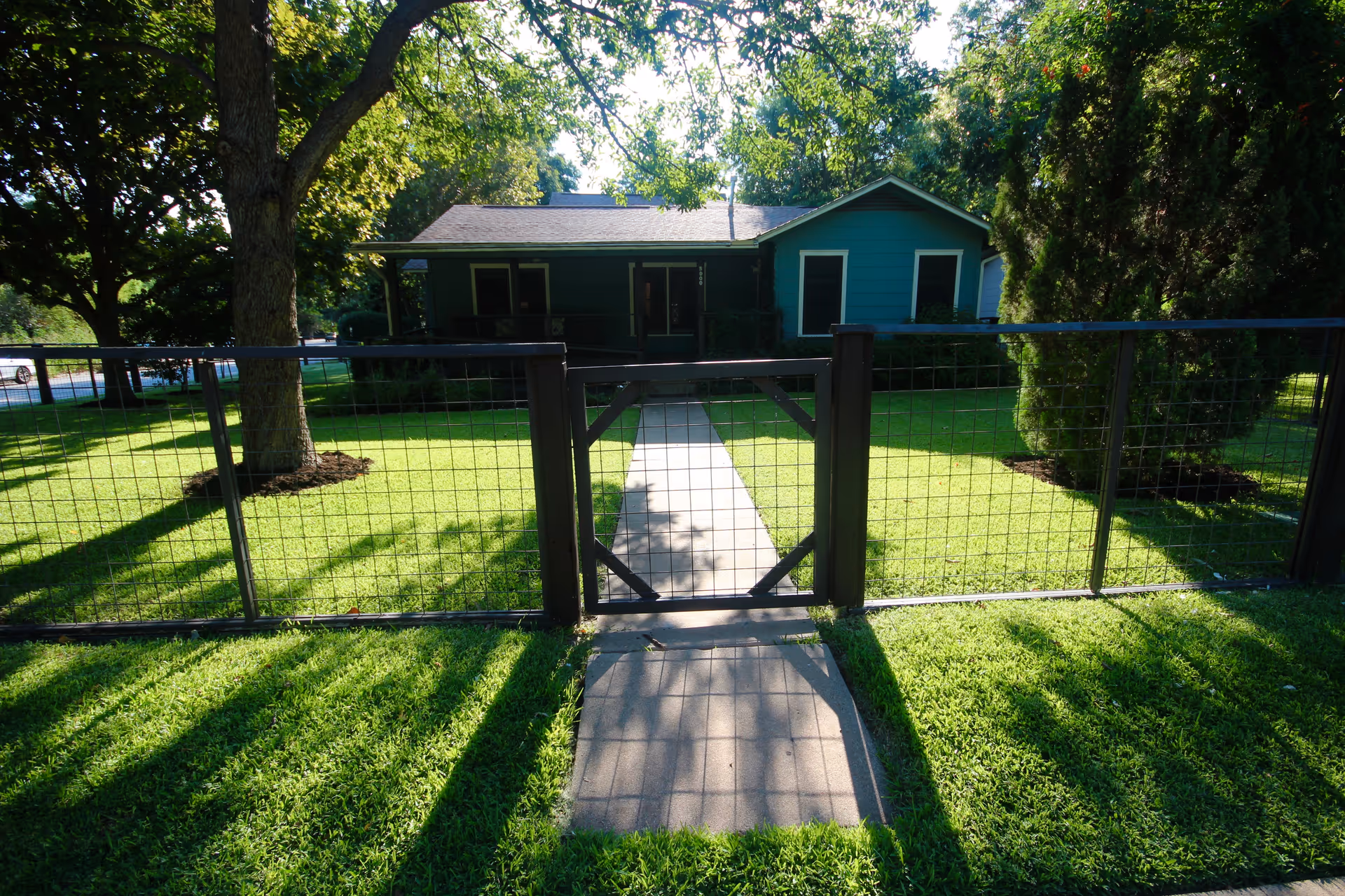 View of a single-story blue house with a front porch, surrounded by a green lawn and trees. A metal fence with a gate encloses the yard, and a concrete pathway leads from the gate to the house entrance.