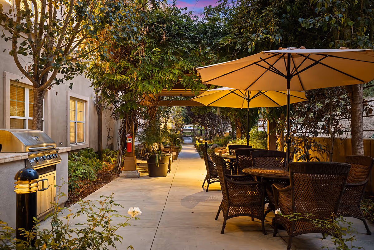 Outdoor patio area with wicker chairs and tables under large beige umbrellas, surrounded by lush greenery and trees. A stainless steel barbecue grill is positioned near the building wall with windows. The pathway is well-lit with warm lighting, leading to a gated entrance in the background.