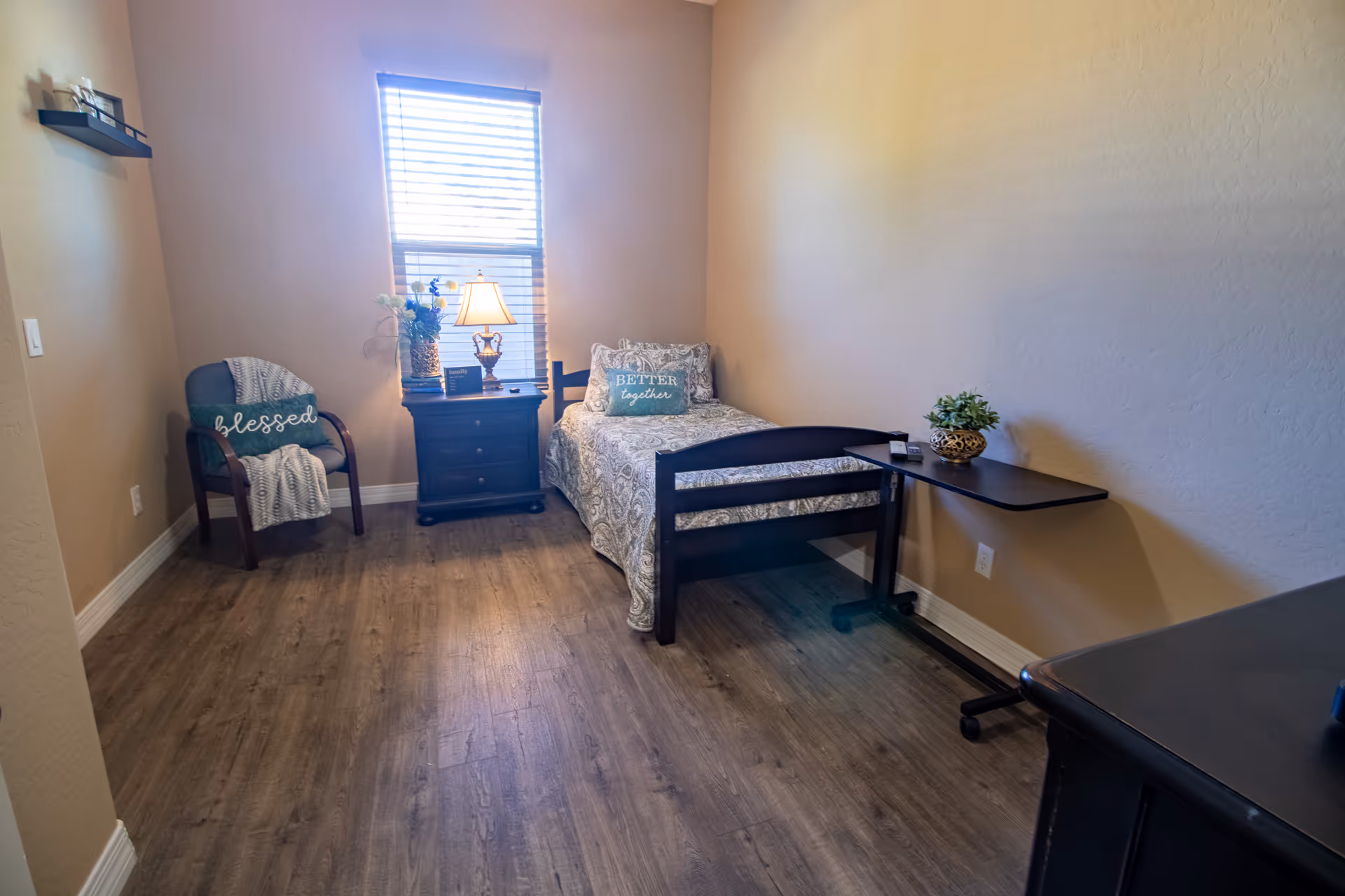 Small bedroom with a single bed, nightstand and lamp by a window, a chair with a 'blessed' pillow, and an overbed table.