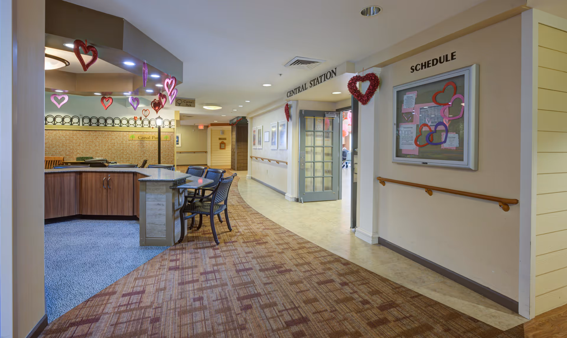 Interior hallway of a senior living facility decorated with hanging heart-shaped ornaments. On the left, there is a counter area with chairs and a sign that reads 'Canal Park.' The hallway has a carpeted floor transitioning to a tiled floor near a door labeled 'Central Station.' On the right wall, there is a bulletin board labeled 'Schedule' with heart decorations.
