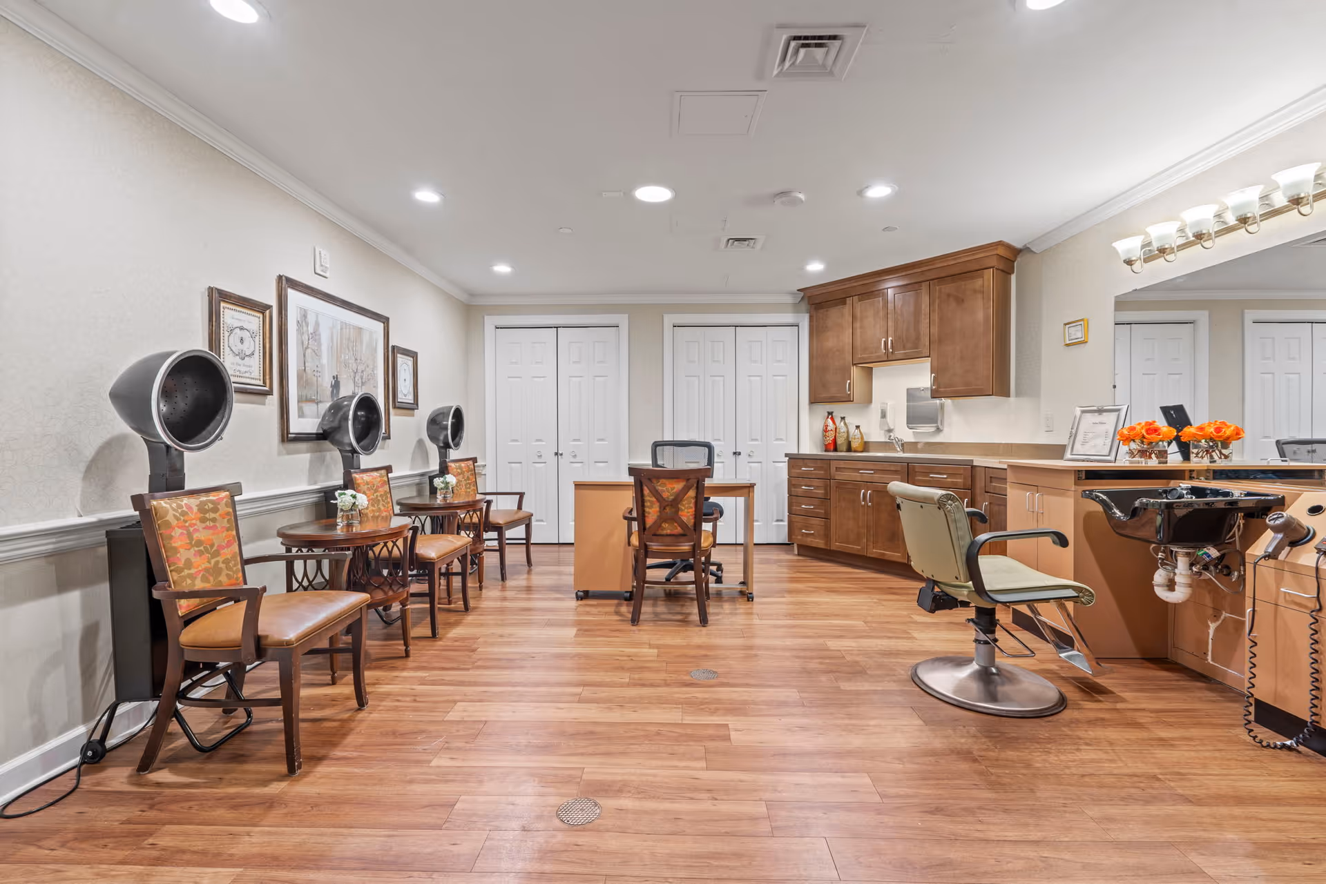 Bright salon/styling room with multiple hooded hair dryers, salon chairs, a styling sink and cabinetry on wood floors.