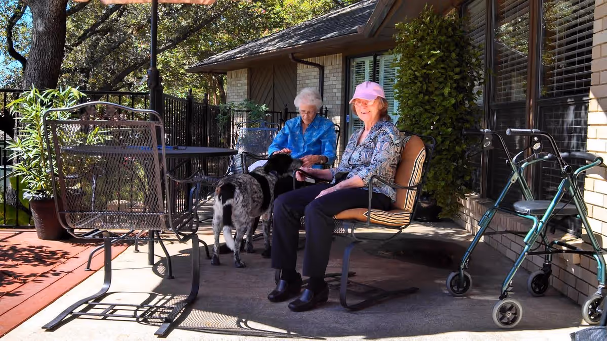 Two elderly women sitting on a patio outside a brick building, one wearing a pink cap and petting a black and white dog, with a walker nearby and metal patio furniture around them under the shade of trees.