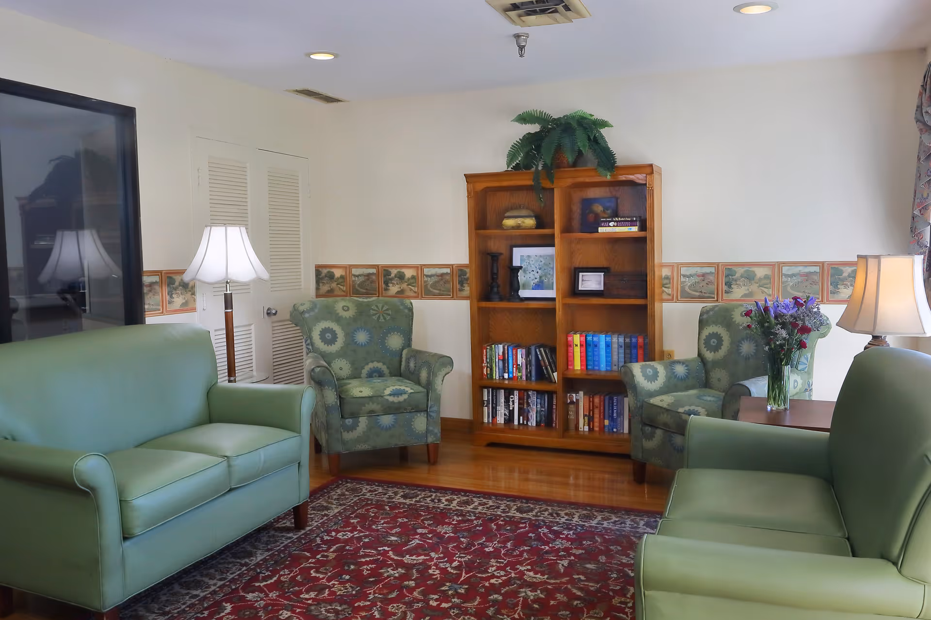 A cozy living room with two green leather sofas and two patterned armchairs arranged around a red patterned rug. There is a wooden bookshelf filled with books and decorative items against the wall, a floor lamp beside one sofa, and a table with a vase of flowers beside one armchair. The walls are white with a decorative border and there is a window with patterned curtains.