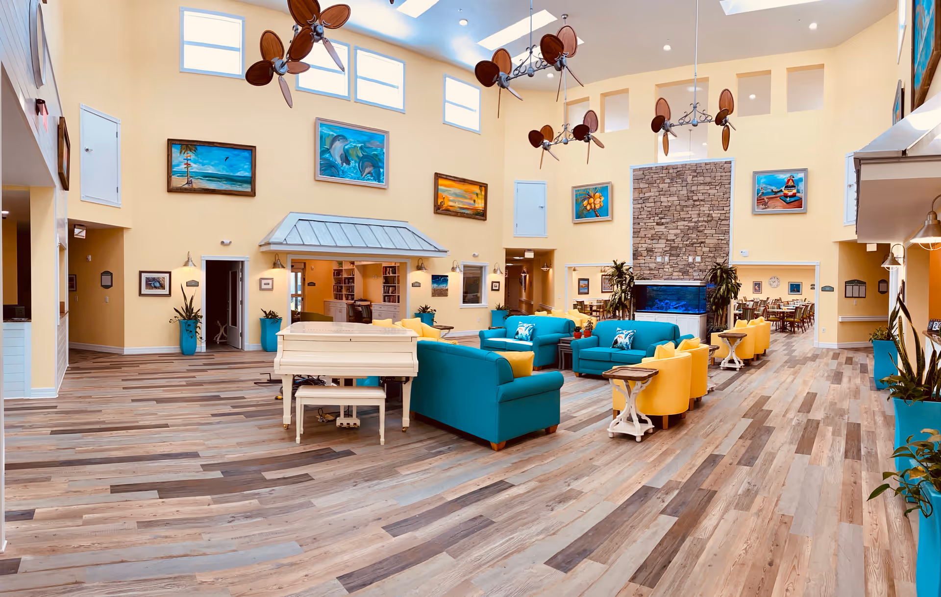 Spacious and brightly lit common area in an assisted living facility with high ceilings, multiple ceiling fans, colorful paintings on the walls, teal and yellow armchairs arranged around a white piano, and a large stone fireplace with an aquarium underneath. The room has wood-patterned flooring and several potted plants.