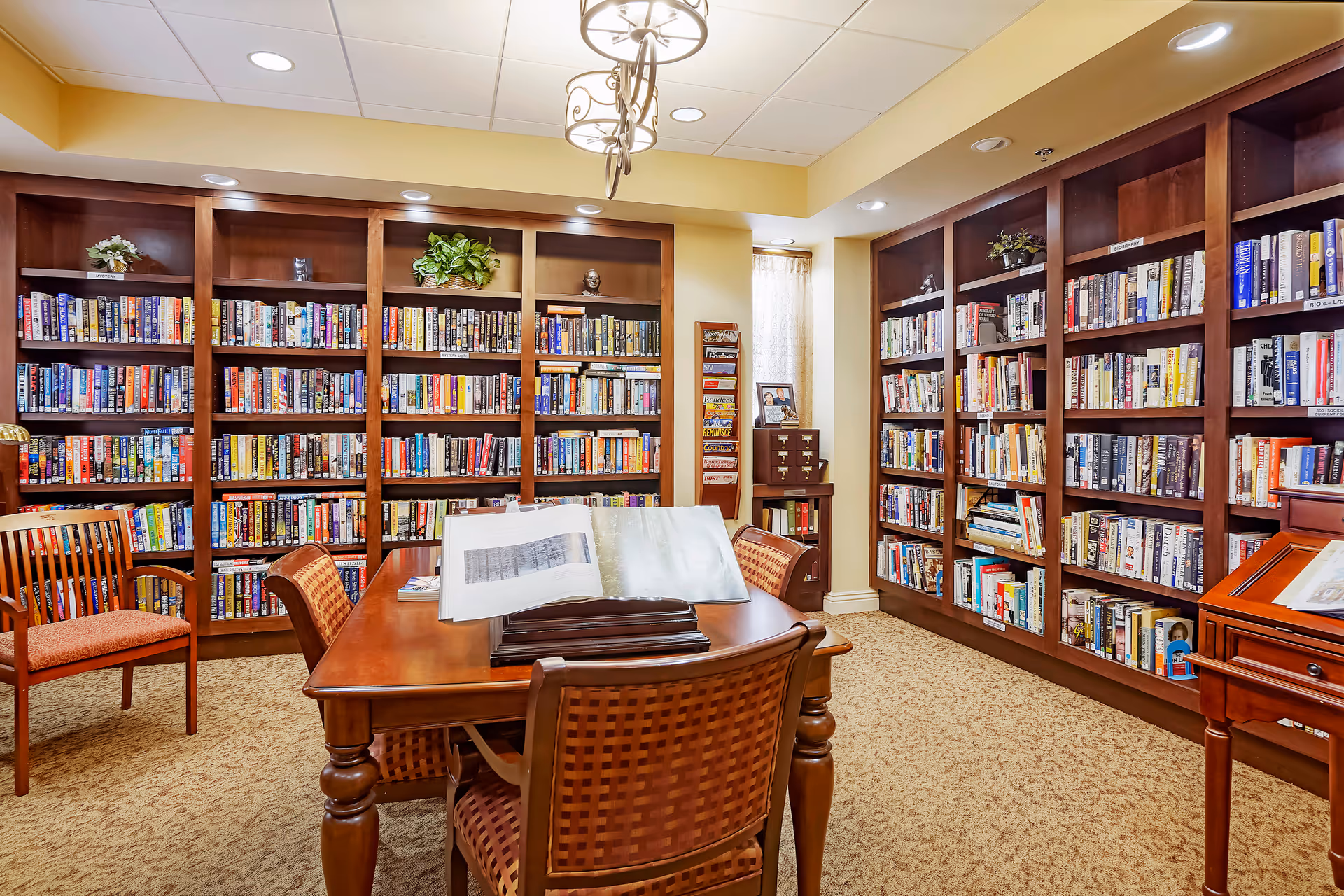 A cozy library room with wooden bookshelves filled with books lining two walls. A wooden table with an open book on a stand is surrounded by four cushioned chairs. The room is warmly lit with ceiling lights and a decorative hanging light fixture. There are small plants and decorative items on top of the bookshelves, and a window with a sheer curtain in the corner.