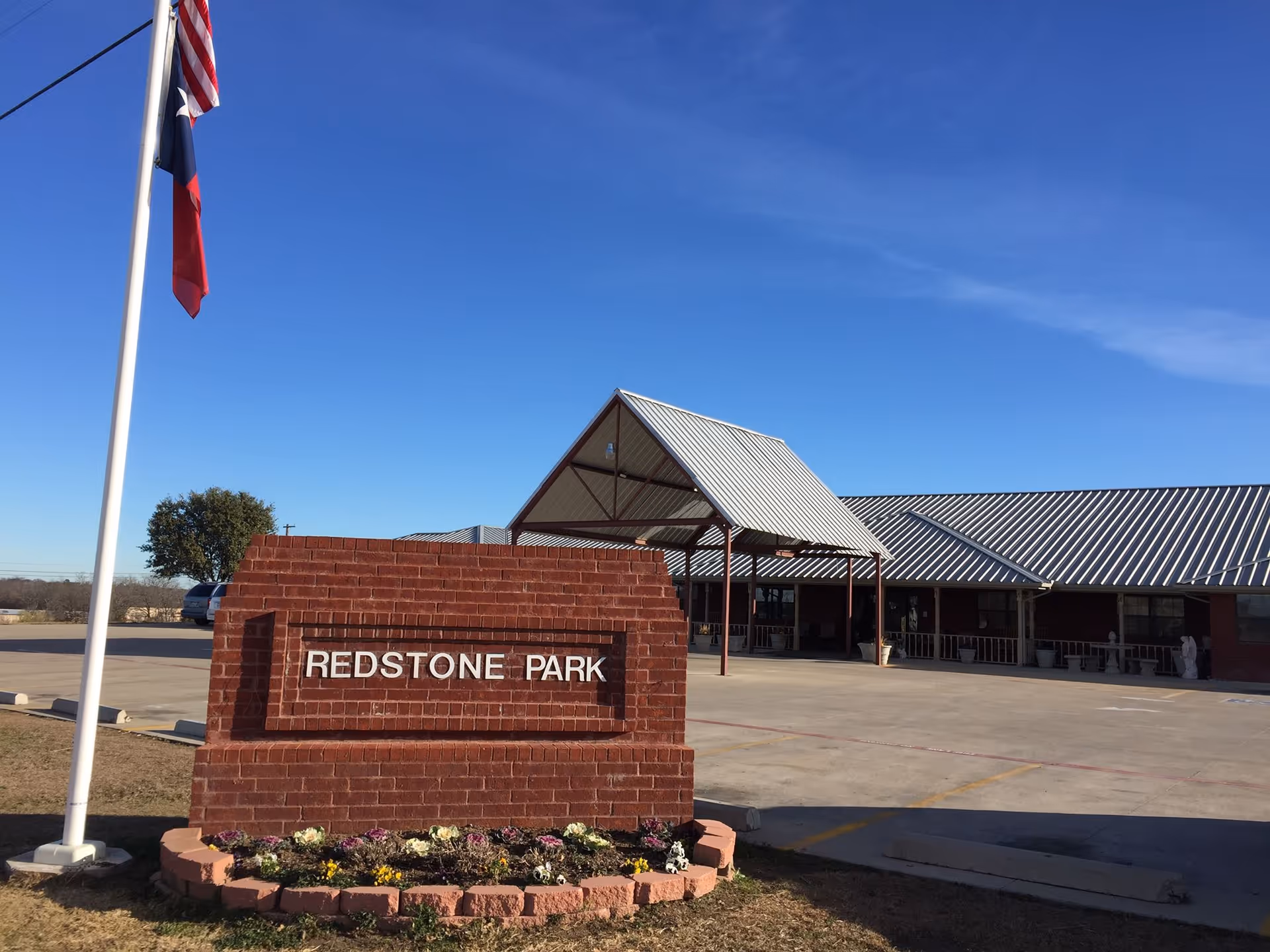Exterior view of the Redstone Park facility showing a brick sign with the facility name, a flagpole with two flags, a parking area, and a building with a metal roof under a clear blue sky.