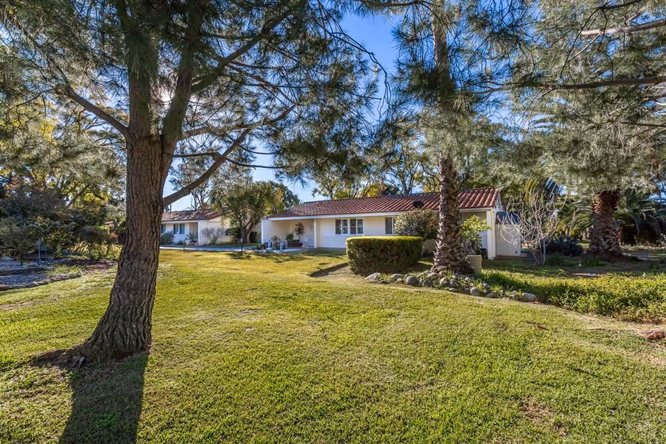 Single-story building with a red-tile roof set behind a grassy lawn and trees in a landscaped yard.