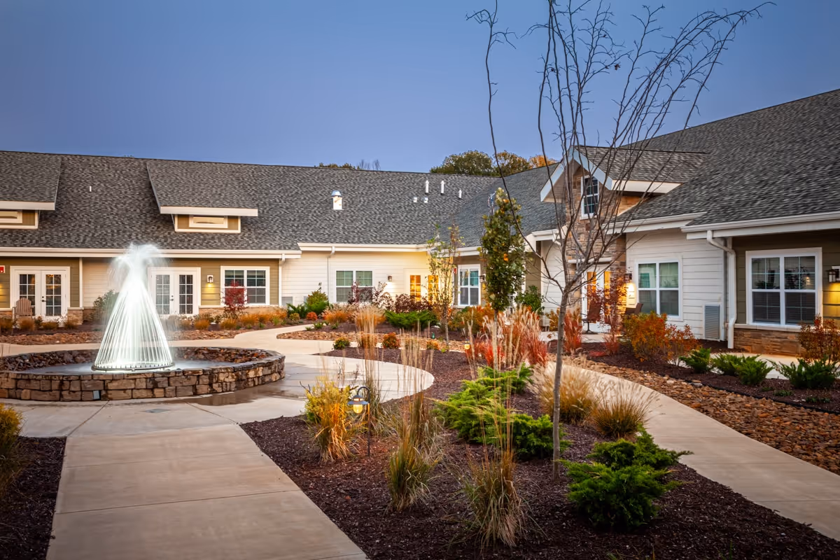 Outdoor courtyard area of a senior living facility with a central water fountain, landscaped garden beds with shrubs and ornamental grasses, and a paved walkway surrounded by single-story buildings with multiple windows and doors under a clear evening sky.