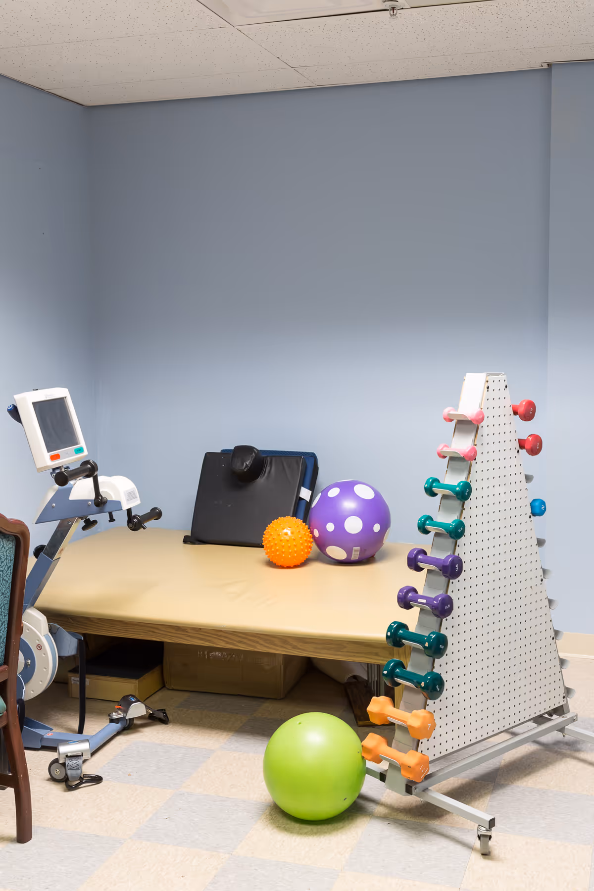 A small rehabilitation room with a beige padded therapy table, colorful therapy balls, a rack holding various small dumbbells, and a piece of exercise equipment with a screen. The walls are painted light blue and the floor has a checkered pattern of beige and gray tiles.