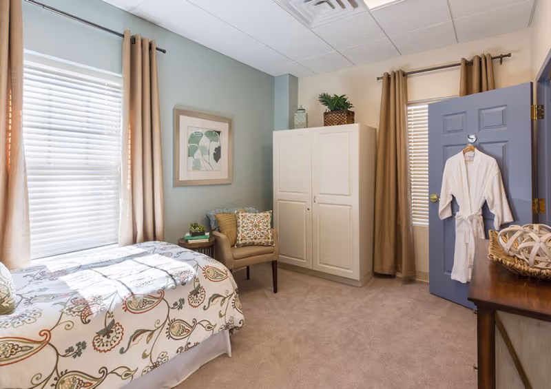 Well-lit furnished bedroom with a single bed, patterned bedding, an armchair, wardrobe, and a robe hanging on the door.