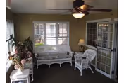 Sunlit sitting room featuring white wicker chairs and a loveseat, ceiling fan, potted plants, and French doors overlooking a courtyard.
