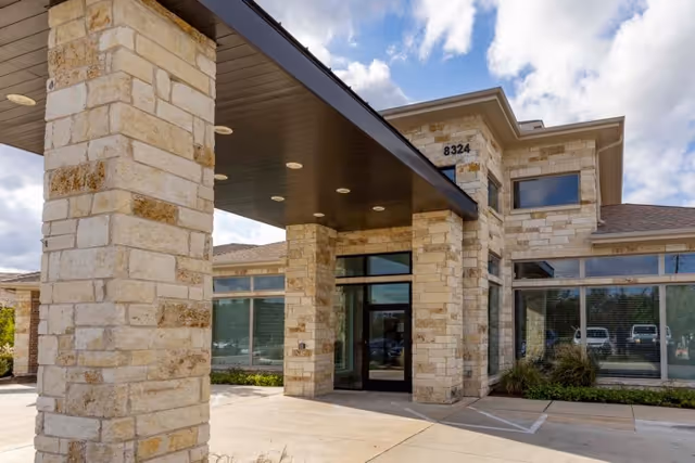 Exterior view of Sedona Trace Health & Wellness building entrance with stone pillars, large windows, and a covered driveway under a partly cloudy sky.