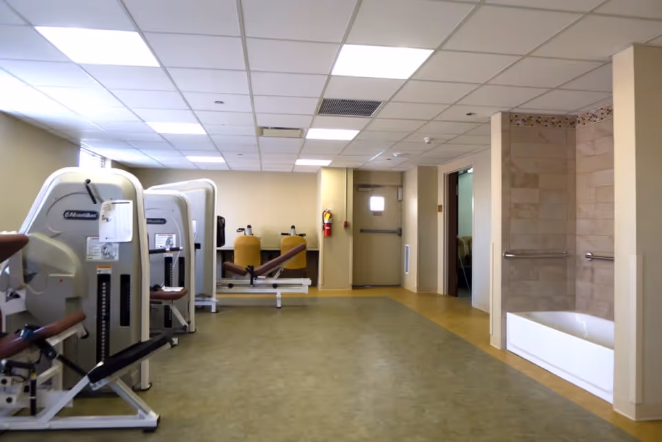 Rehabilitation room with exercise machines and a walk-in bathtub/shower area in a senior living facility.