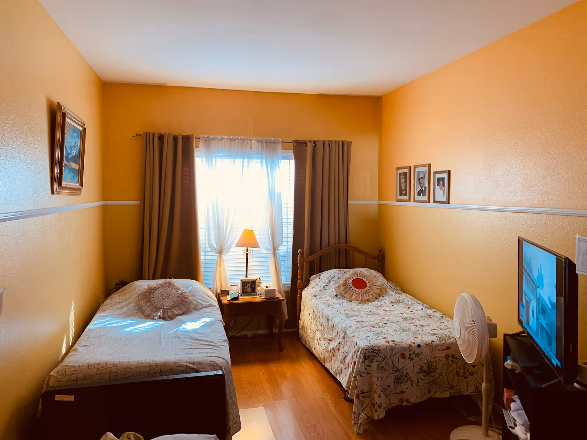 A cozy bedroom with two single beds separated by a wooden nightstand with a lamp and picture frames. The room has warm yellow walls, a window with sheer white curtains and brown drapes, a standing fan near a TV on a small stand, and framed pictures on the walls.