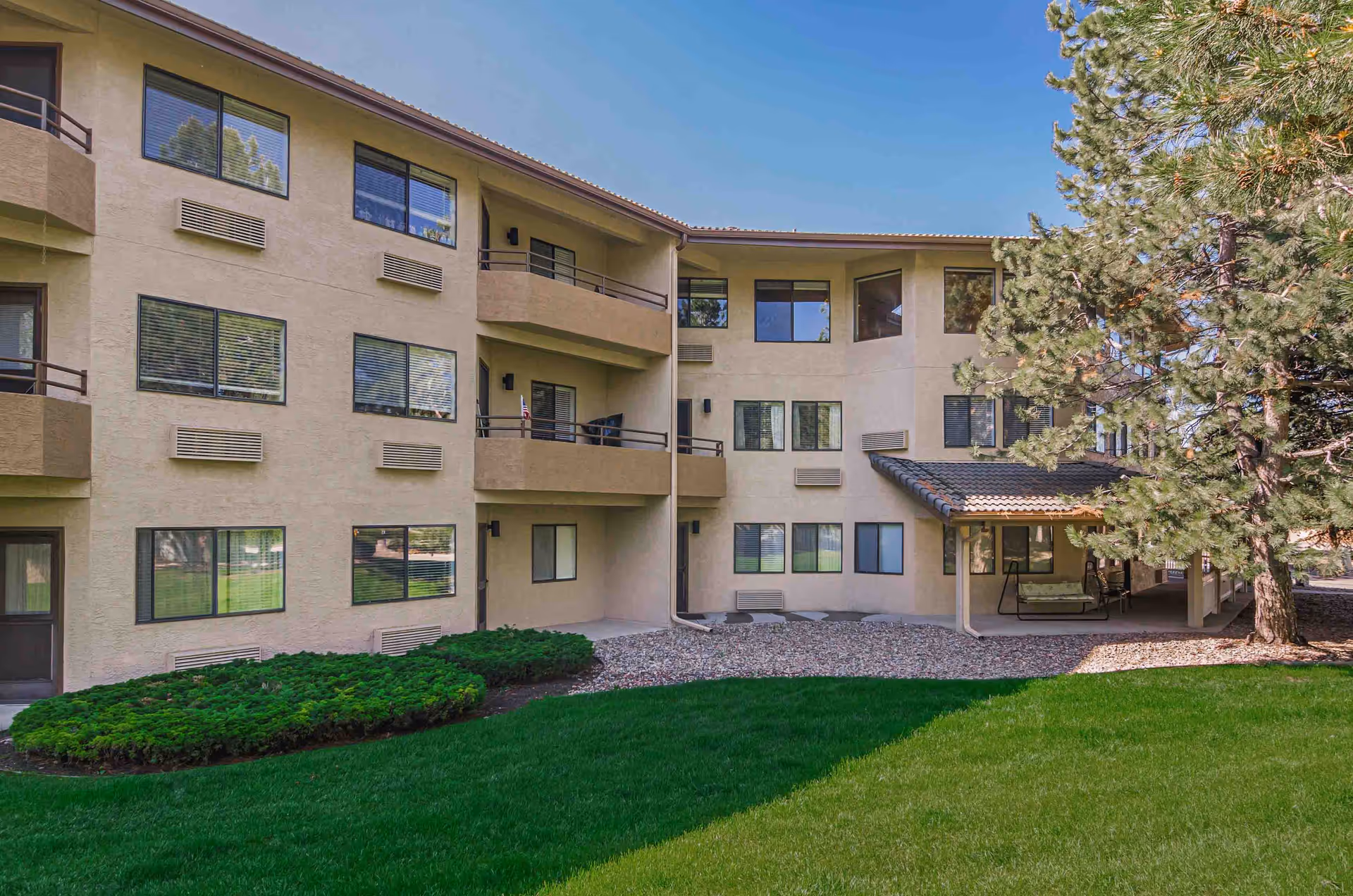 Exterior view of a beige multi-story residential building with balconies, windows, and a covered patio area. The building is surrounded by green grass, bushes, and tall pine trees under a clear blue sky.