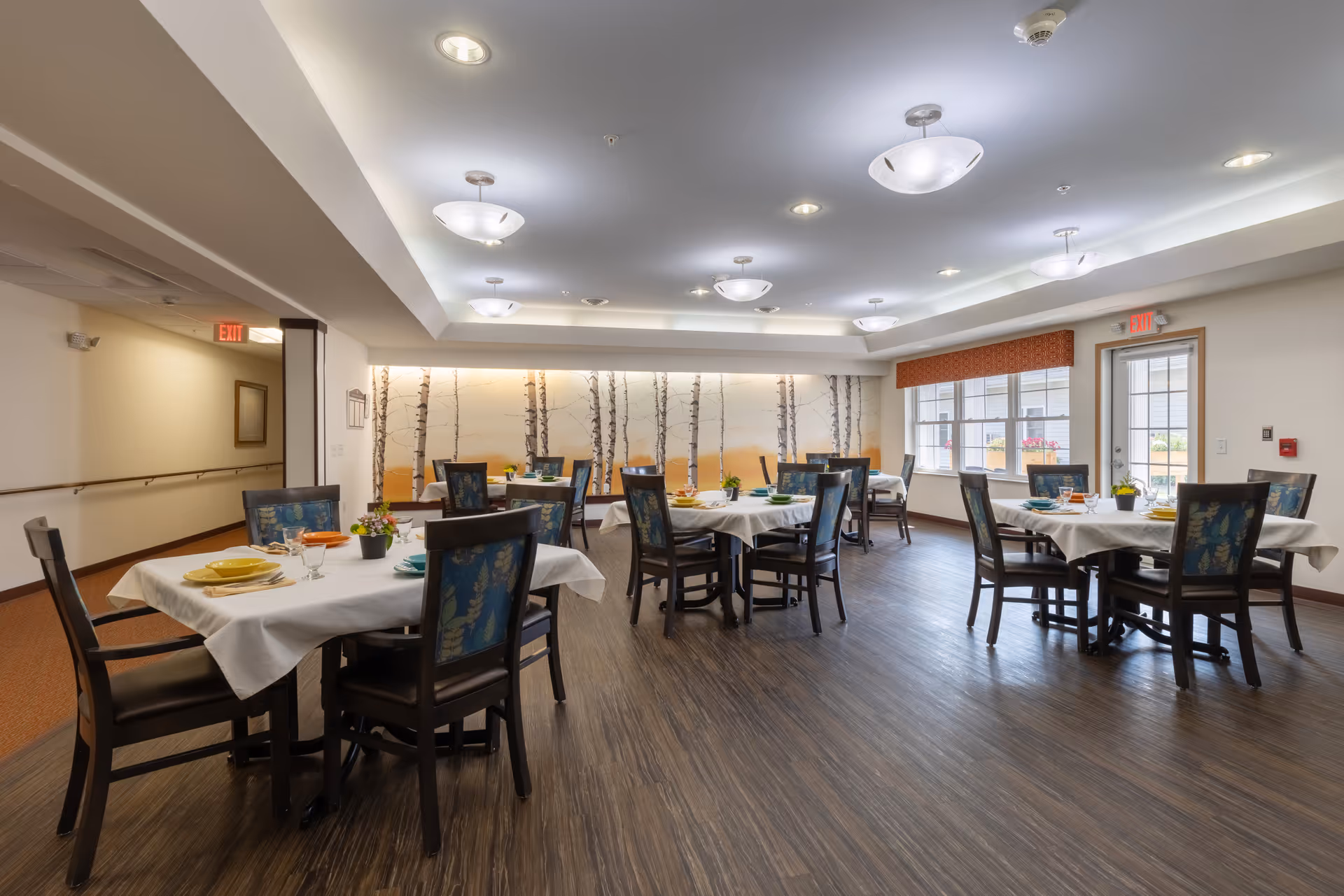 A bright dining room in a senior living facility with multiple round tables covered with white tablecloths. Each table is set with colorful plates, glasses, and small flower arrangements. The room has wood flooring, modern ceiling lights, large windows with an orange valance, and a wall mural featuring birch trees.