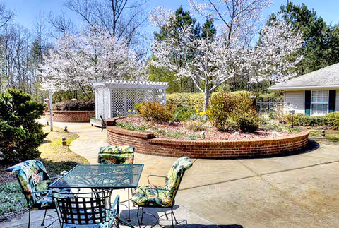 Sunlit outdoor courtyard with a patio table and chairs, raised brick planter, blooming trees and a white pergola beside a senior living building.
