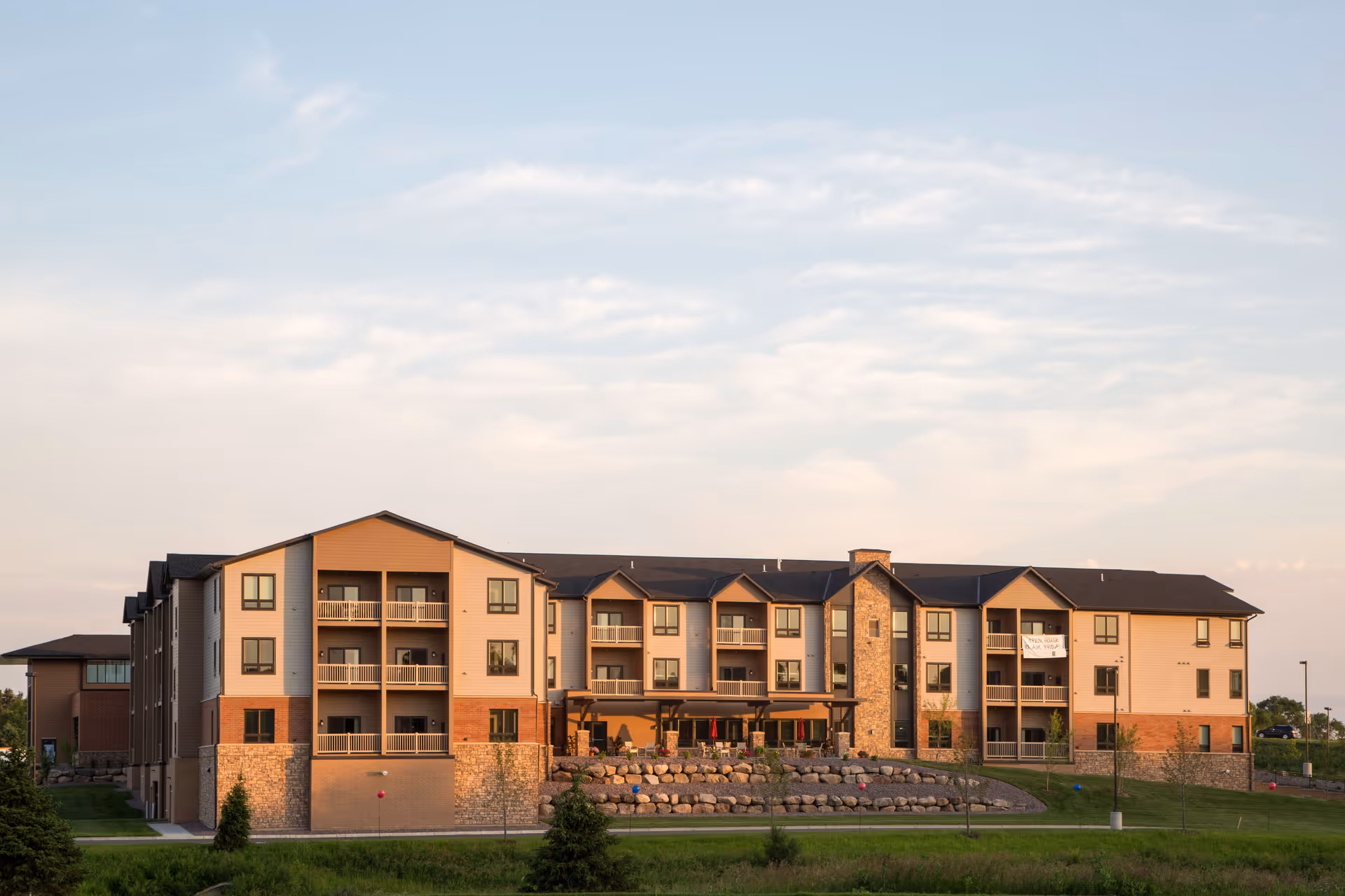 Exterior view of a three-story senior living facility building with balconies, stone and brick facade, and a grassy area in front under a partly cloudy sky.