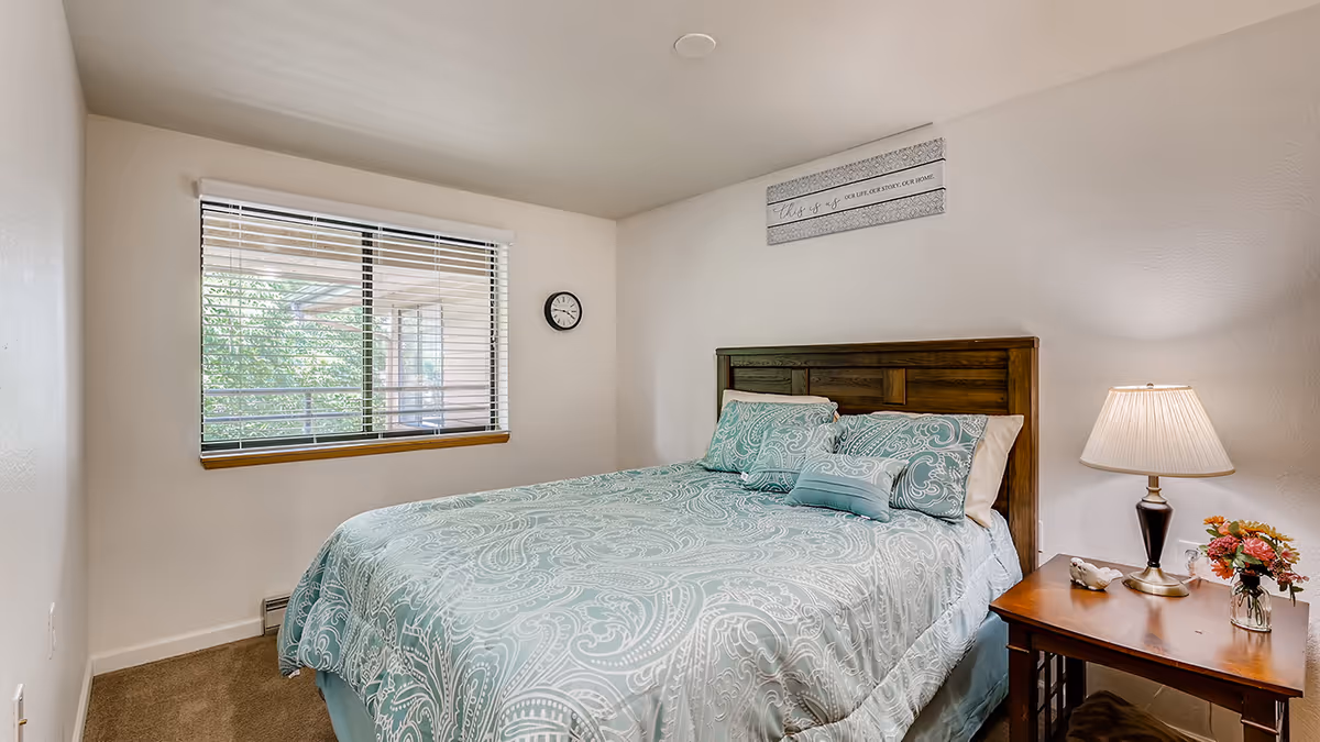 A cozy bedroom with a large bed covered in a light blue and white patterned comforter and matching pillows. There is a wooden headboard, a wooden nightstand with a lamp, a small vase of flowers, and a decorative bird figurine. A window with blinds lets in natural light, and a small clock is mounted on the wall next to the window. Above the bed is a wall decoration with text.